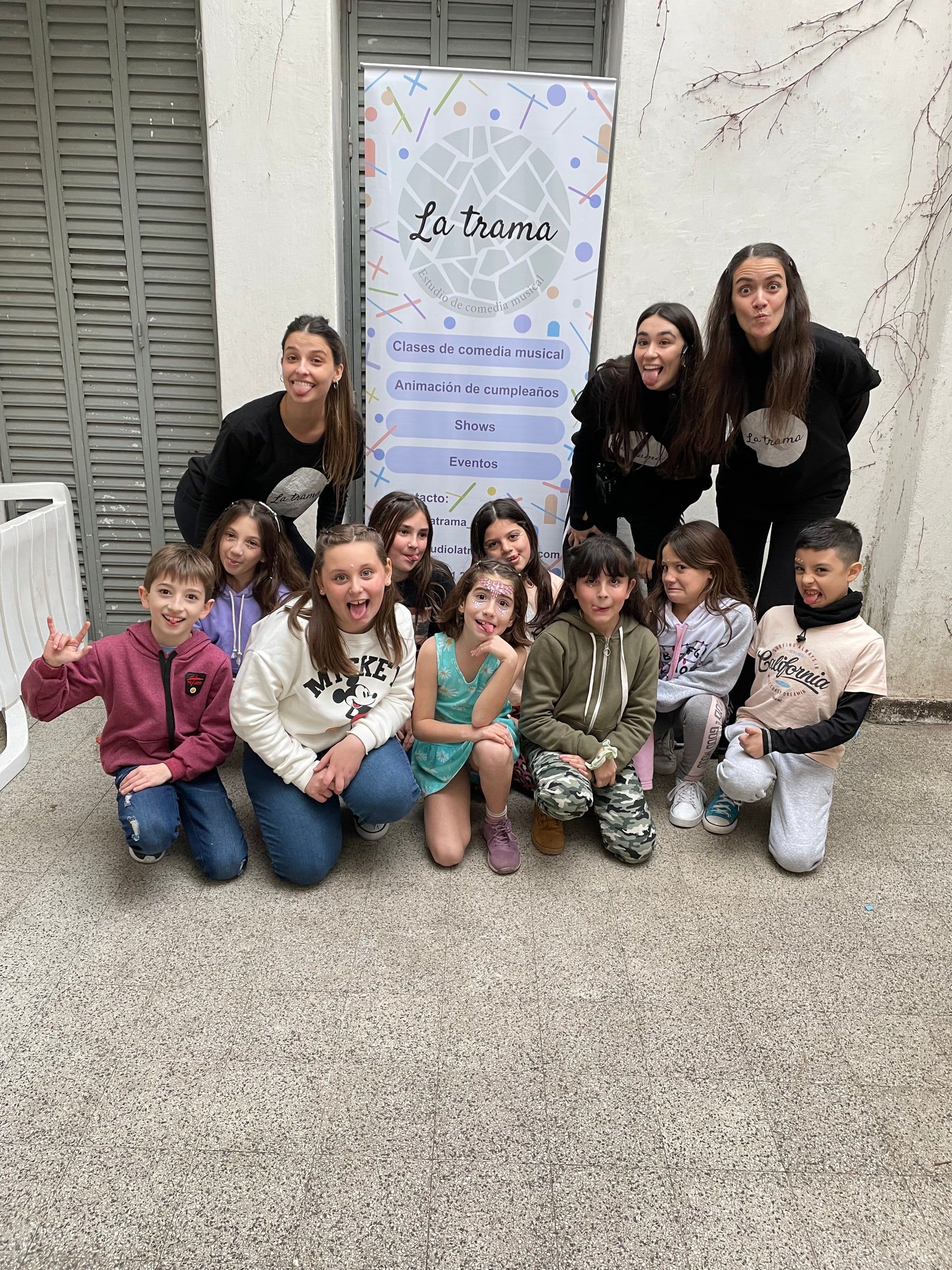 Grupo de chicos y chicas sentados en el piso del estudio junto a dos adultas, delante de un cartel de La Trama.