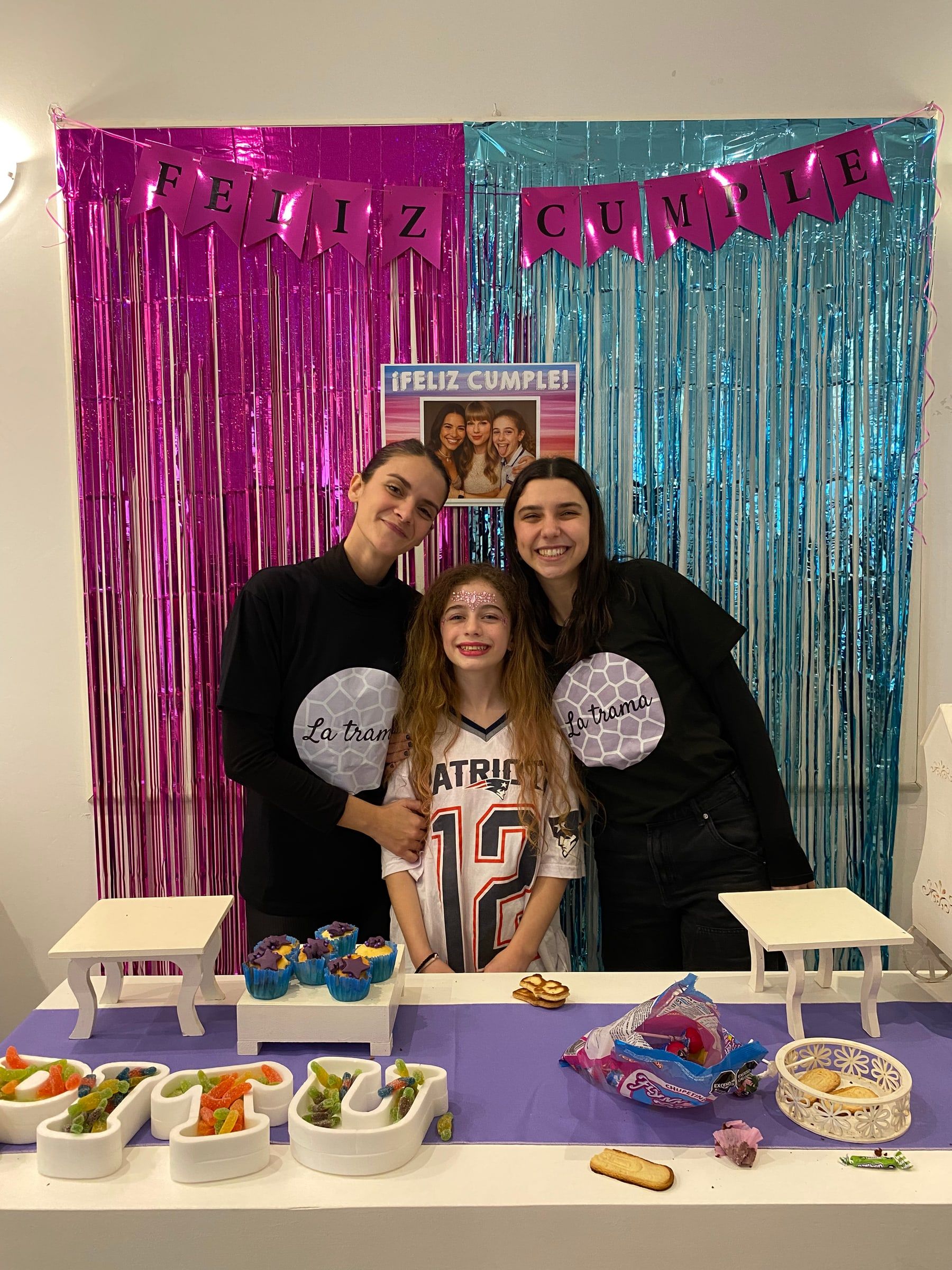 Cumpleañera con dos chicas de La Trama frente a una mesa decorada y una cortina de flecos de colores.