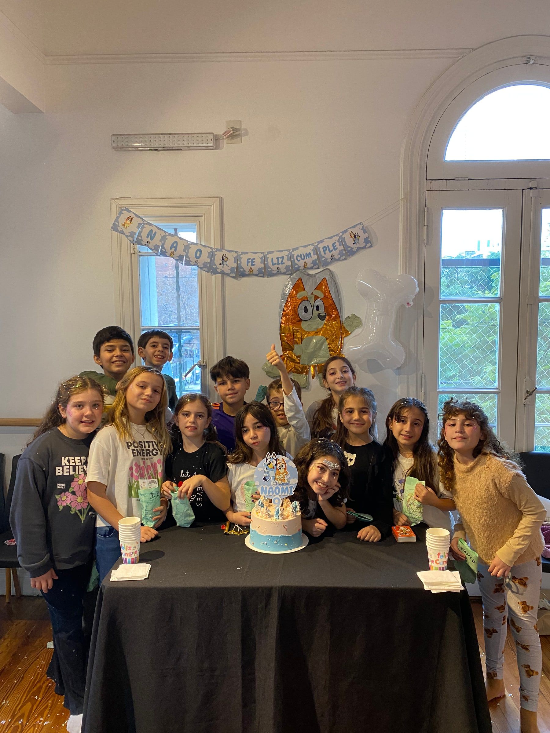 Chicos y chicas alrededor de la mesa con la torta de cumpleaños, en una sala del estudio decorada con globos.