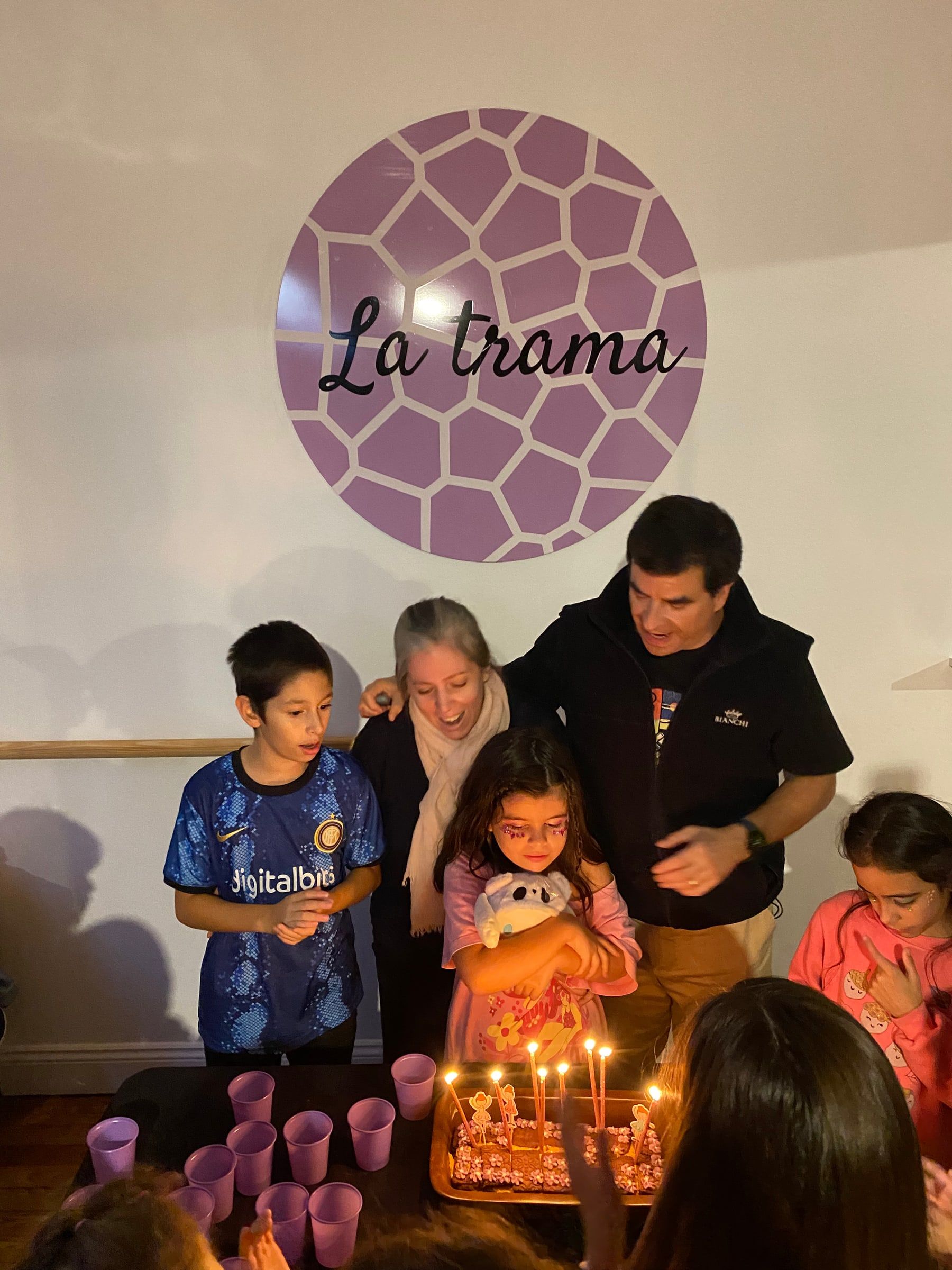 Una cumpleañera sopla las velas rodeada de amigos, con el logo de La Trama en la pared.