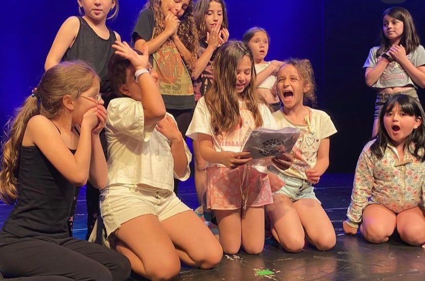 Un grupo de chicas sobre el escenario, sorprendidas, actuando una escena con un papel en la mano.