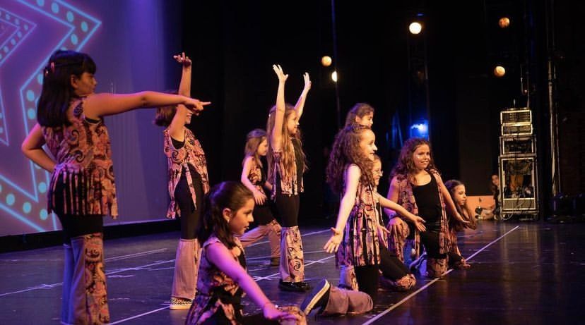 Chicas bailando una coreografía con vestuario estampado en un escenario con luces violetas.