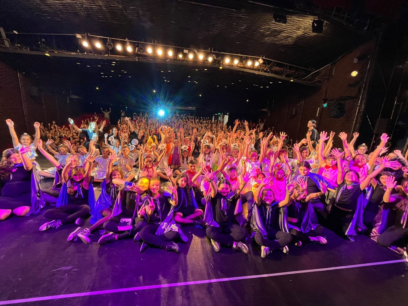 Alumnas y alumnos de La Trama saludan desde el escenario con los brazos en alto, bajo las luces de un teatro.