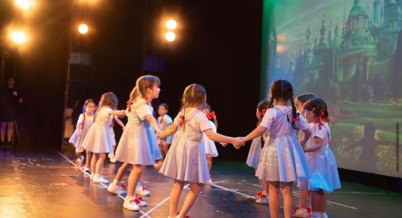 Mini Peques con vestidos blancos bailando en ronda sobre el escenario.
